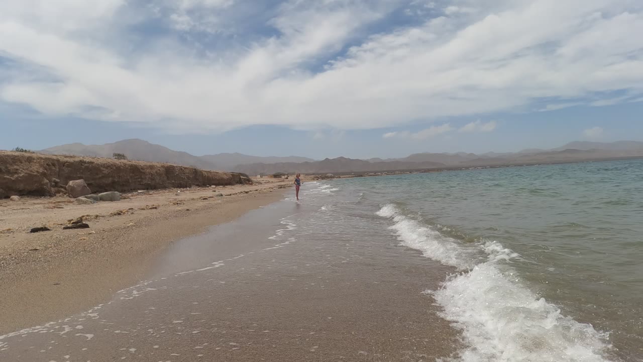 Baja California, Mexico beach at 0:02 with a brighter patch of white foam breaking in the right foreground, one distant person near center, and a hazy mountain range across the horizon.