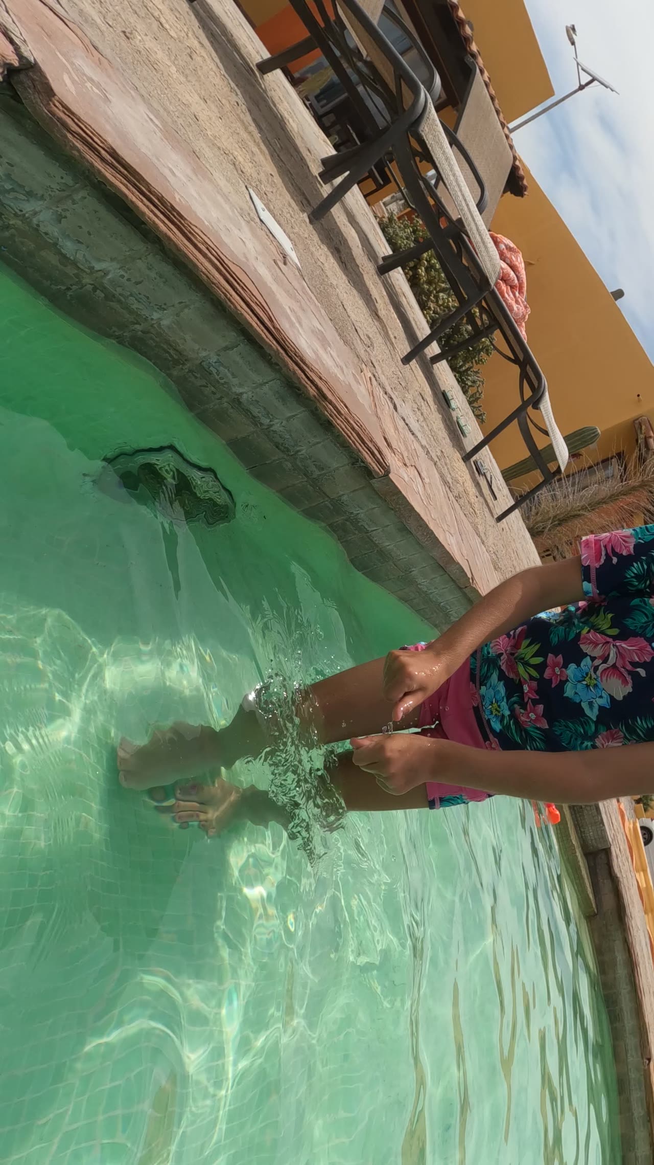 Child standing in shallow pool water with one hand splashing near the pool edge.