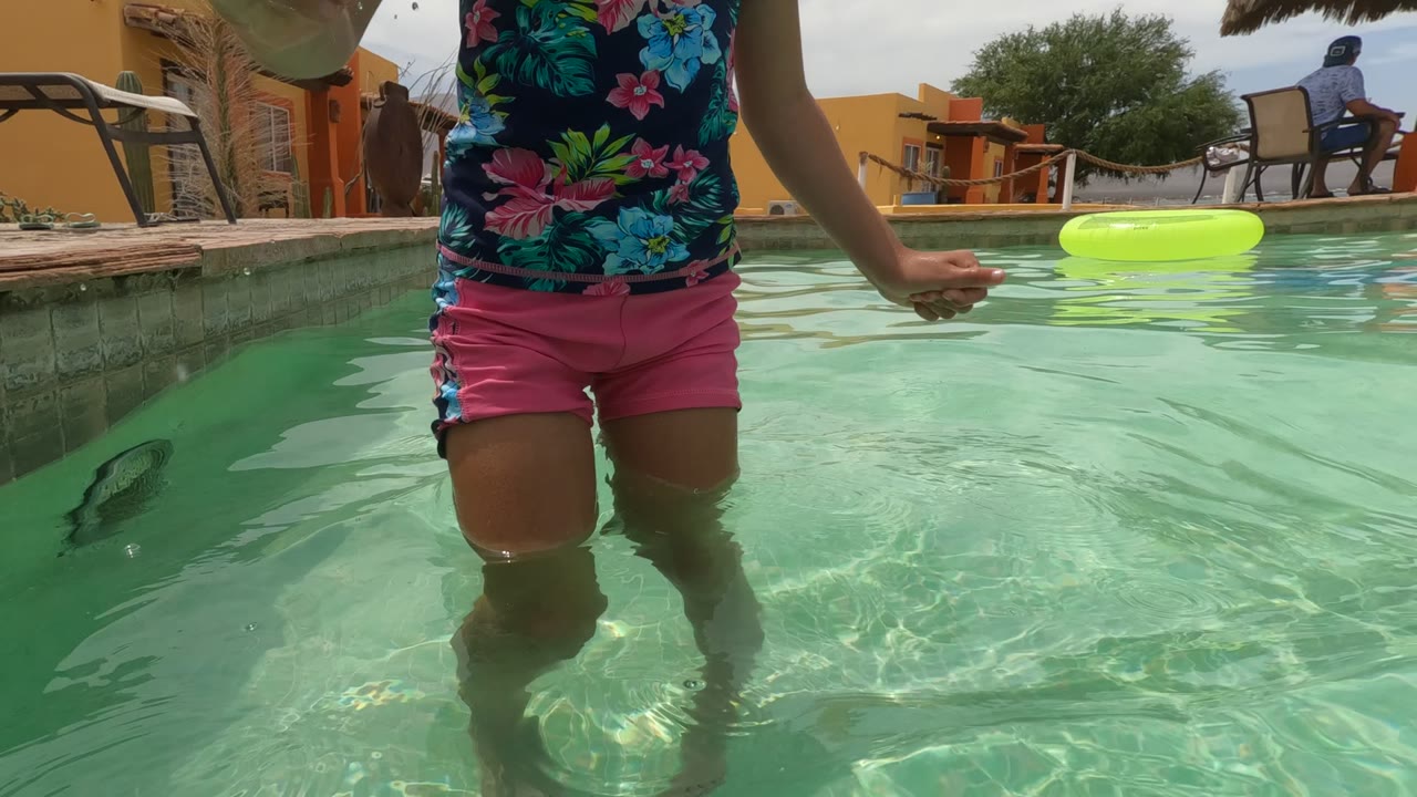 In Baja California, Mexico, the frame crops the child's torso and legs at water level, with rippled clear pool water, the green float, a thatched umbrella, and orange poolside structure behind.