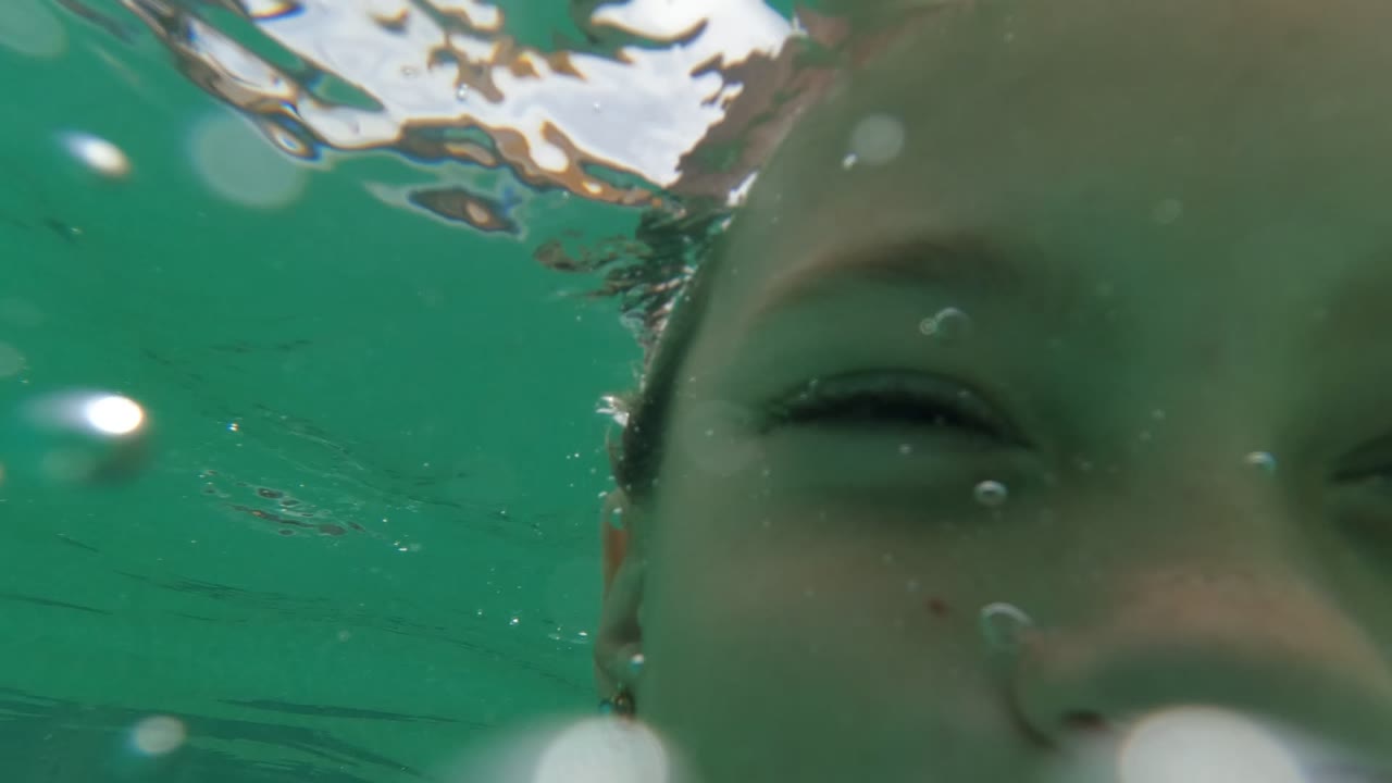 Child's face partially submerged close to the lens with bubbles in green-blue water.