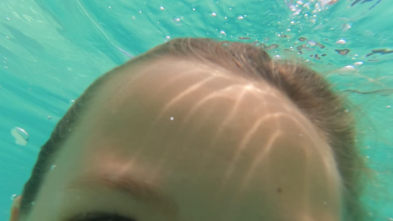 Underwater close-up of the upper part of a child's face in Baja California, Mexico, with bright caustic light patterns and teal pool water around the head.