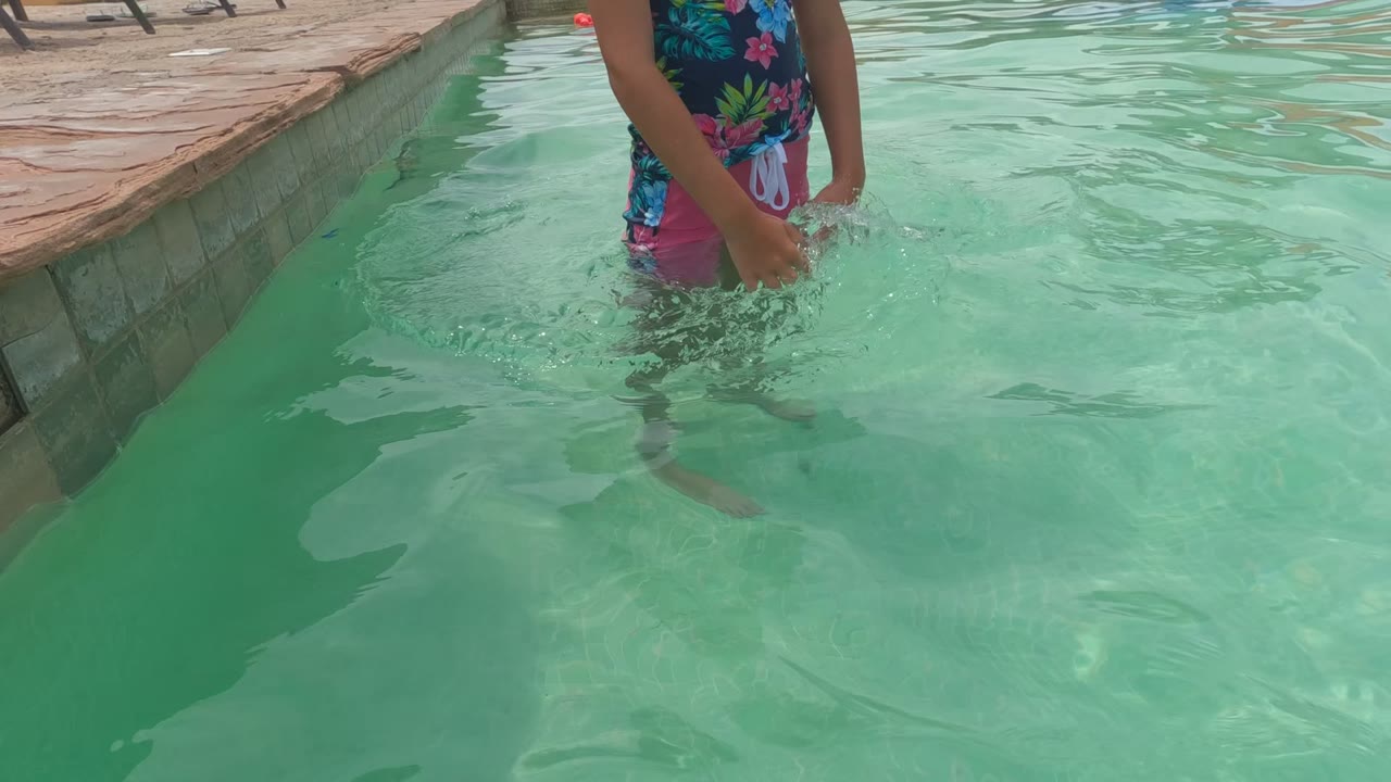 A child in a floral swim shirt stands waist-deep in a Baja California swimming pool, cropped at the upper body with pale green water and the pool edge filling most of the frame.
