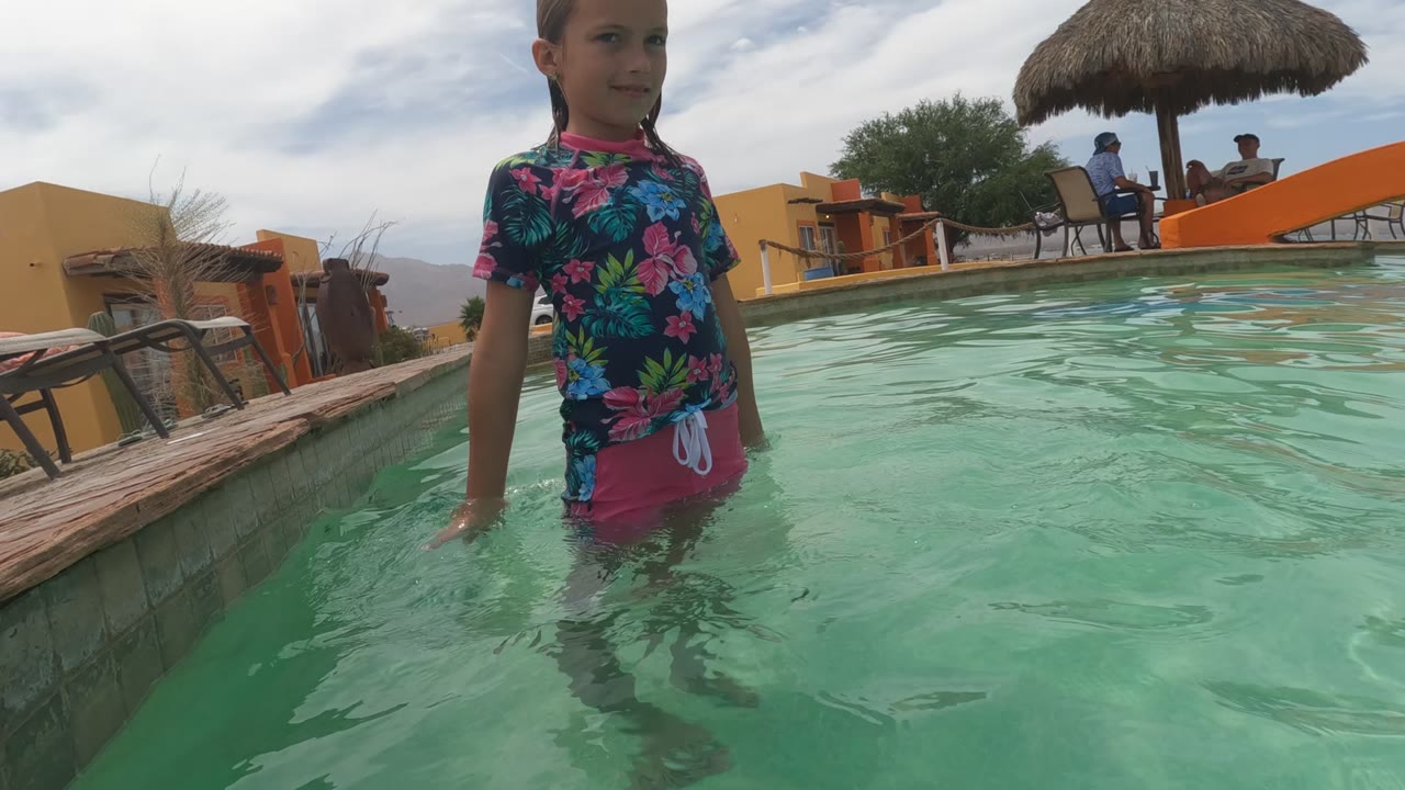 Child in pool with lounge chairs and thatched umbrella behind.