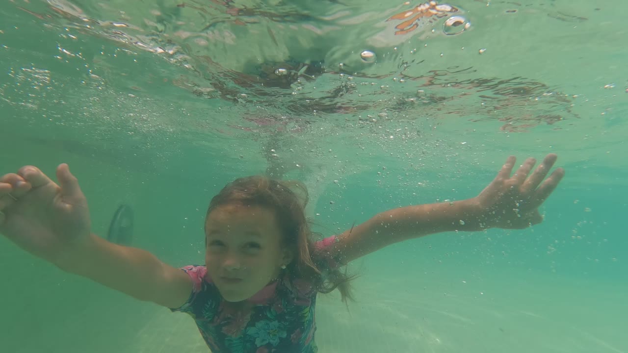 In Baja California, Mexico, a child in a floral swimsuit swims underwater toward the camera with arms spread beneath the rippled pool surface.