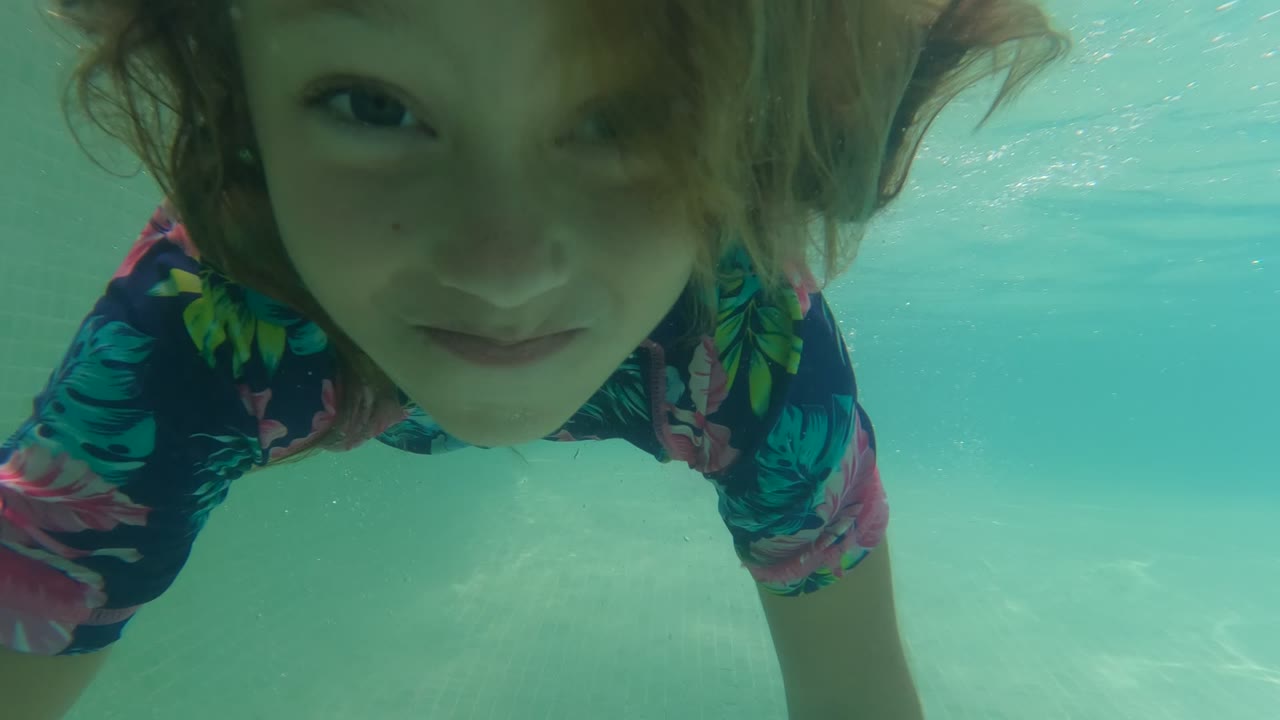 In Baja California, Mexico, the child's face nears the right side of the underwater frame, with the floral swimsuit and turquoise pool water visible around them.