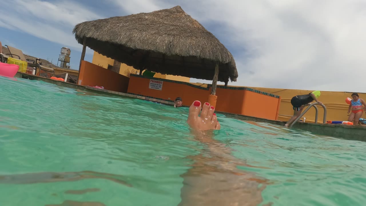 In Baja California, Mexico, feet with red toenails break the surface of a turquoise swimming pool, with a pool edge, ladder, thatched umbrella, small sign, and distant poolside people in the background.
