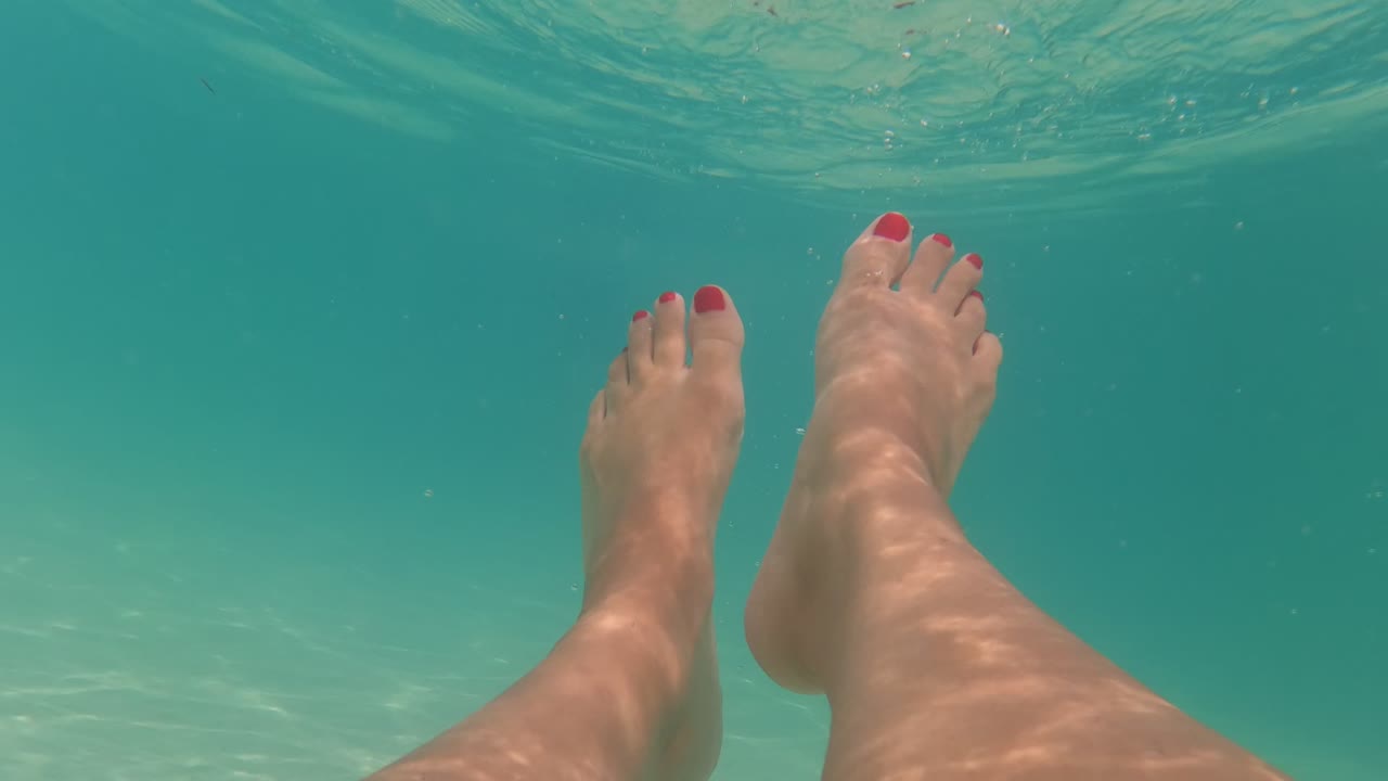 Underwater view of feet with red toenails in clear turquoise pool water.