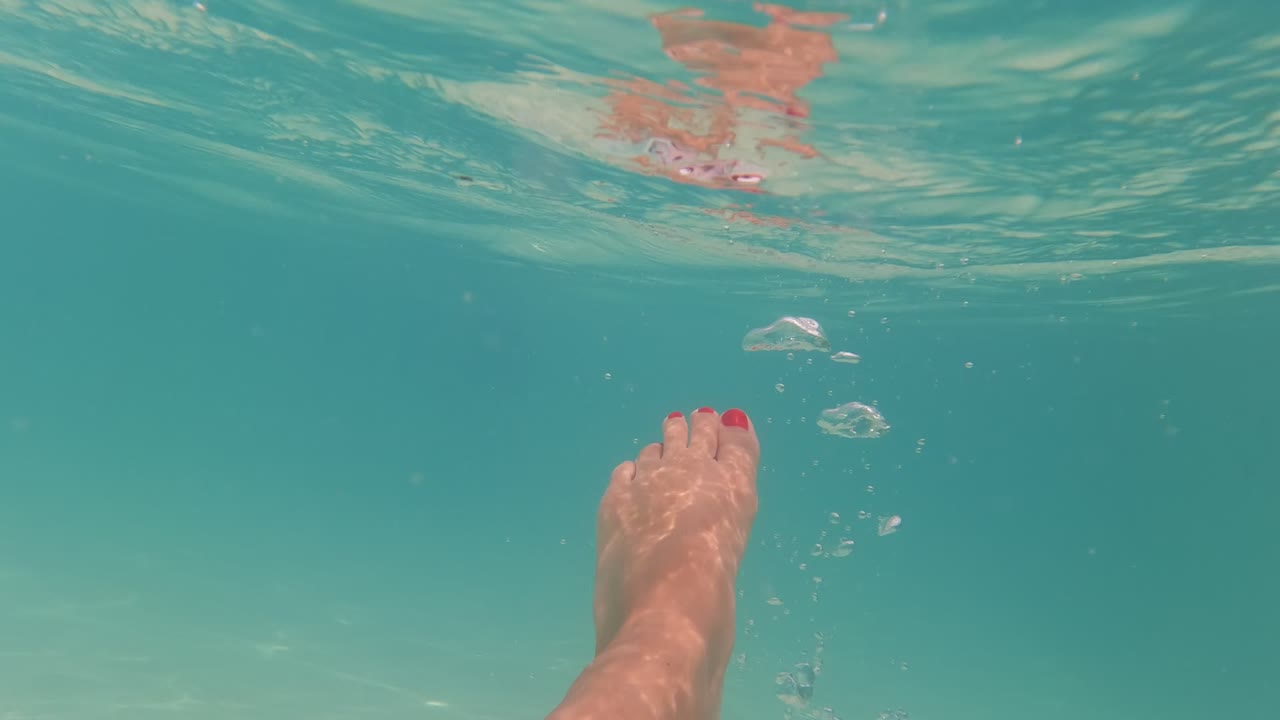 In Baja California, Mexico, a single foot with red toenails angles through the right side of the underwater frame, with small bubbles rising beside it and wavering reflections along the pool surface.