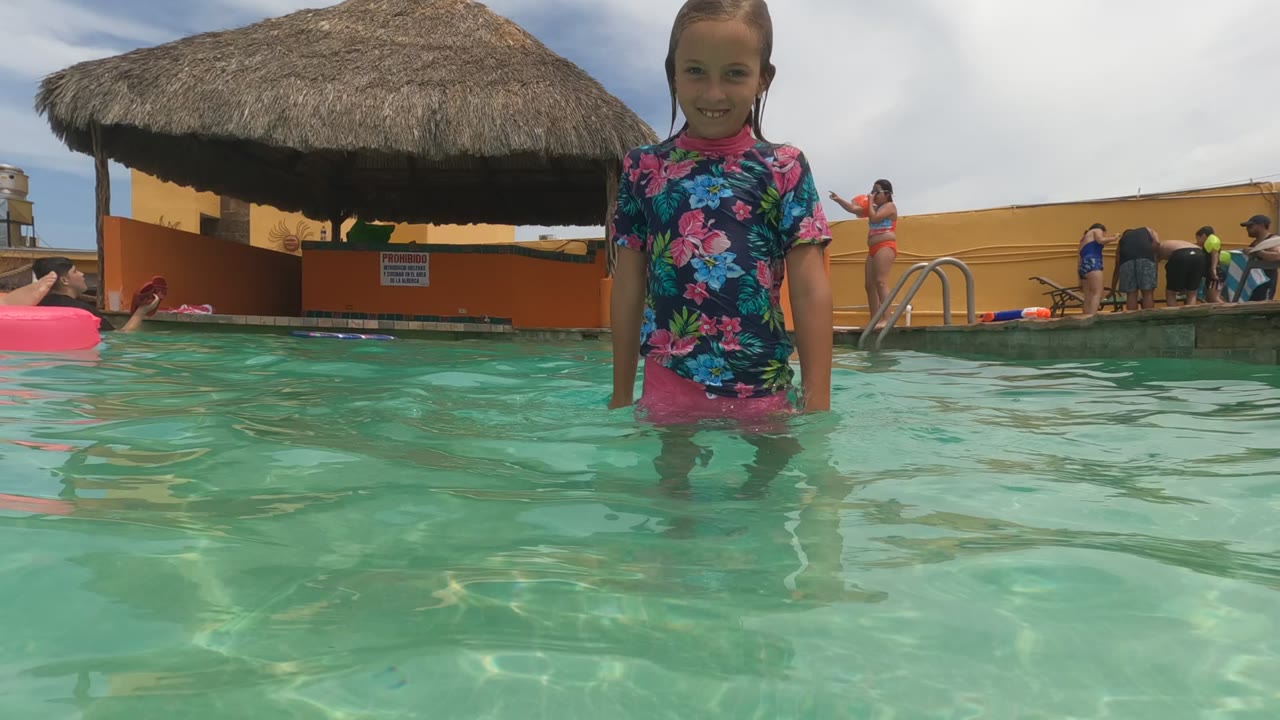 In Baja California, Mexico, a young girl in a floral swim shirt and pink shorts stands in turquoise pool water with a thatched shade roof and poolside people behind her.