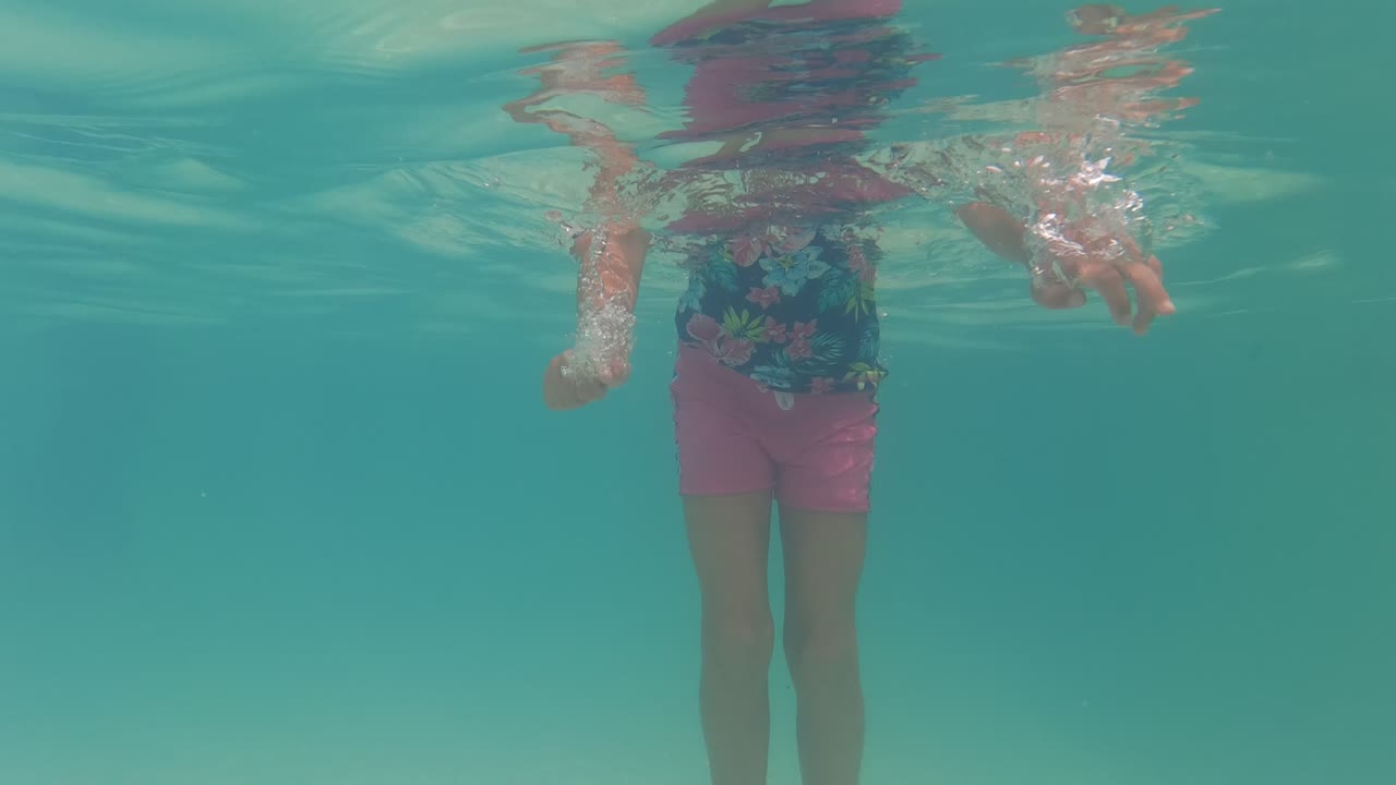 In Baja California, Mexico, the girl floats underwater near the pool surface, with her floral swim shirt, pink shorts, and small bubbles visible in the turquoise water.