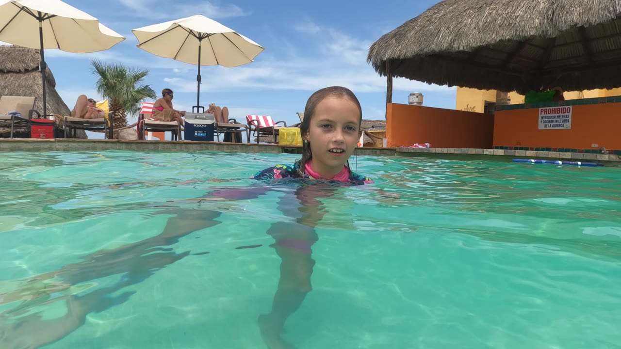 Young girl in Baja California, Mexico at pool surface level, wearing a floral swim shirt as umbrellas, loungers, and a thatched shade roof sit in the sunny background.