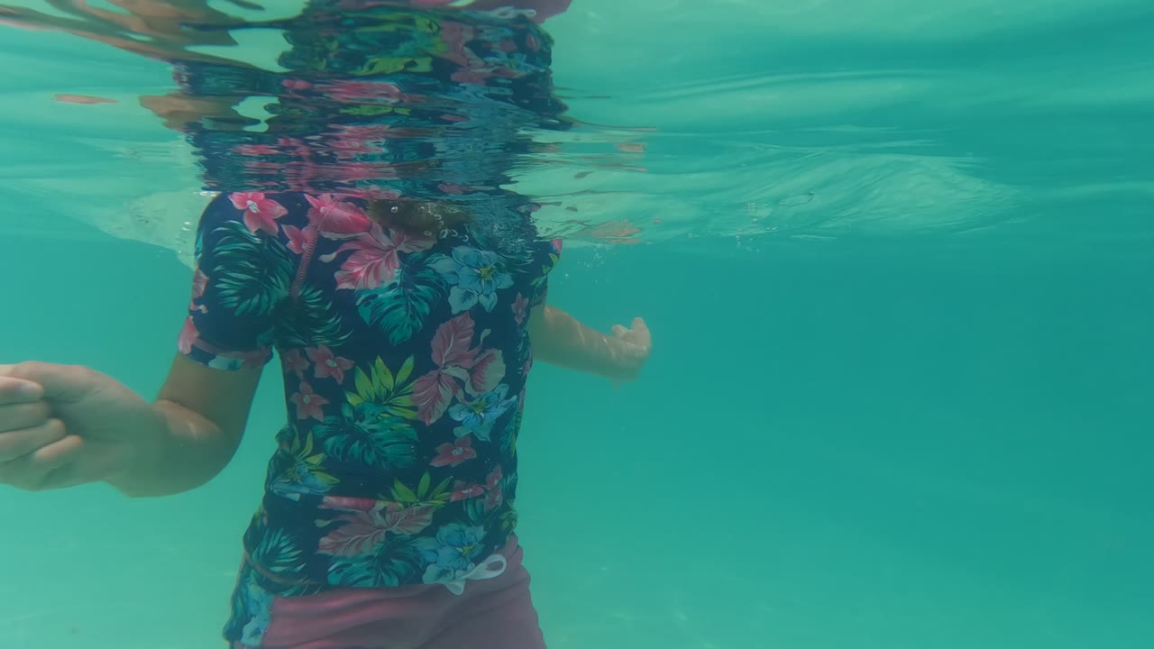 Child underwater in a floral shirt with arms partly raised in turquoise pool water.