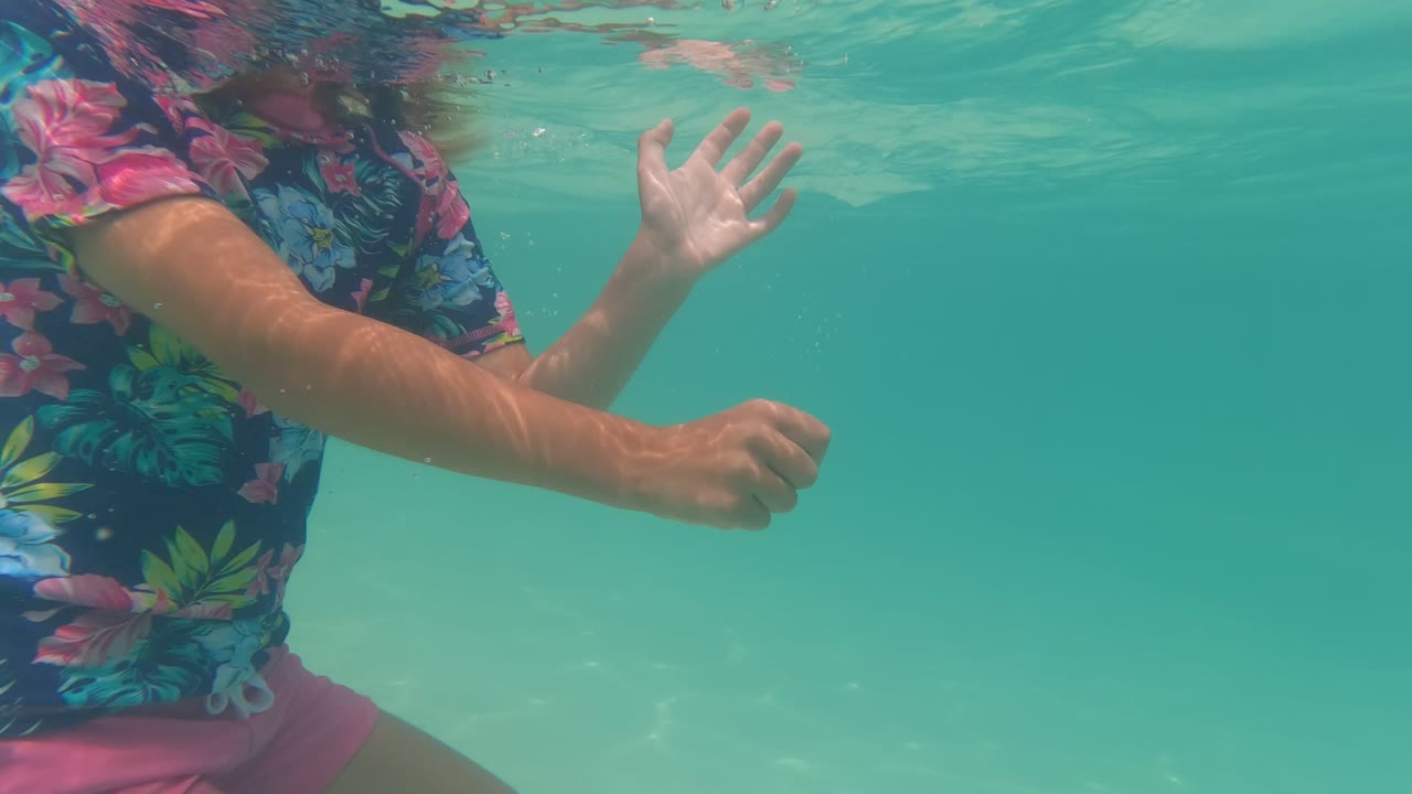 Side view underwater in Baja California, Mexico, showing the child's floral shirt, pink shorts, arms, and hand near the rippled pool surface.