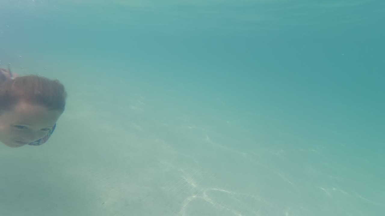 At 0:02 in Baja California, Mexico, the child's face appears near the upper left of a mostly open underwater view, with soft light patterns on the sand below.
