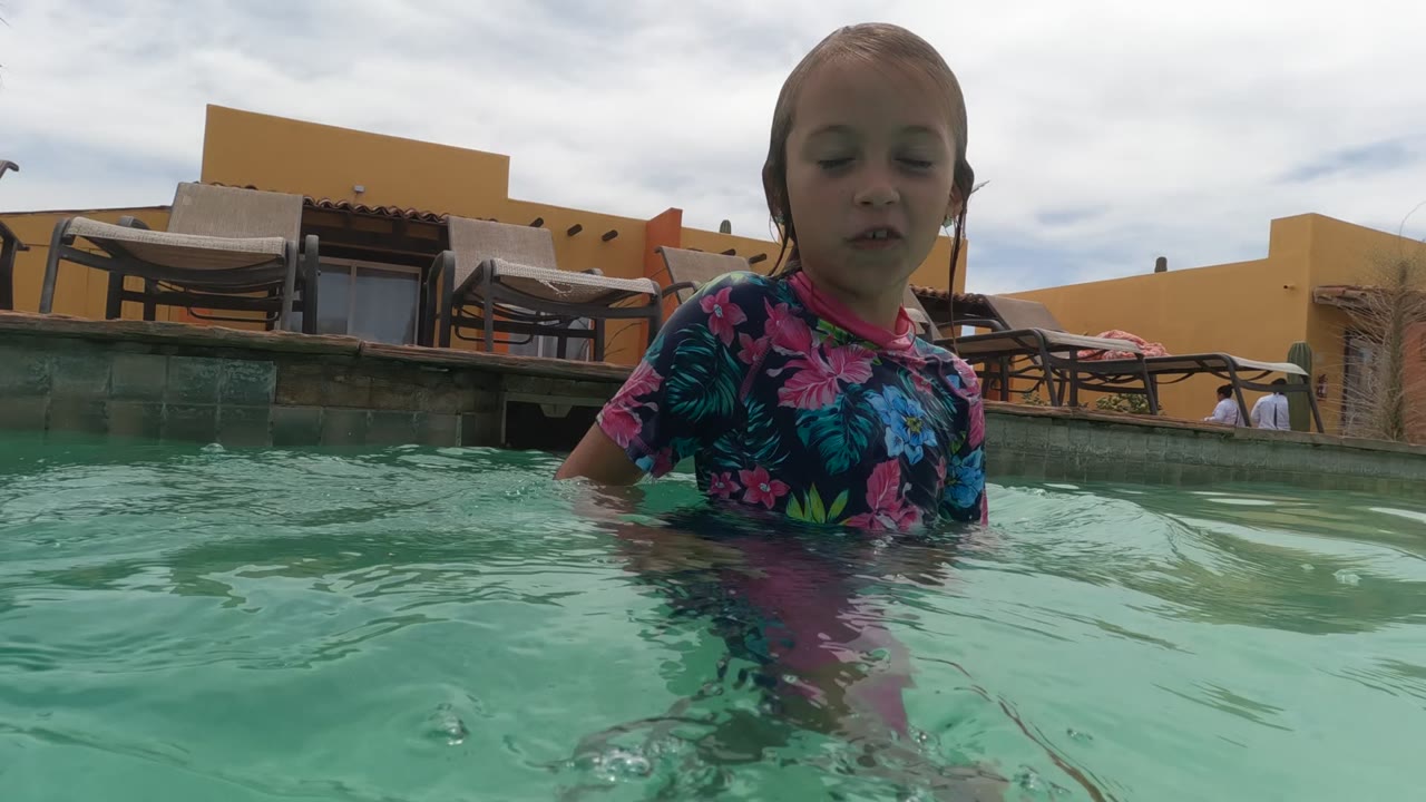 A child in a floral swim shirt stands close to the water-level camera in a Baja California, Mexico swimming pool, with lounge chairs and yellow-orange buildings behind them.