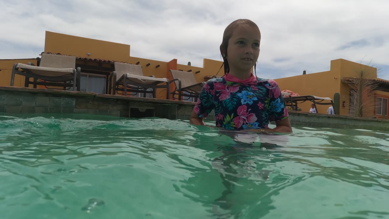 Child standing farther back in rippling pool water with chairs behind.