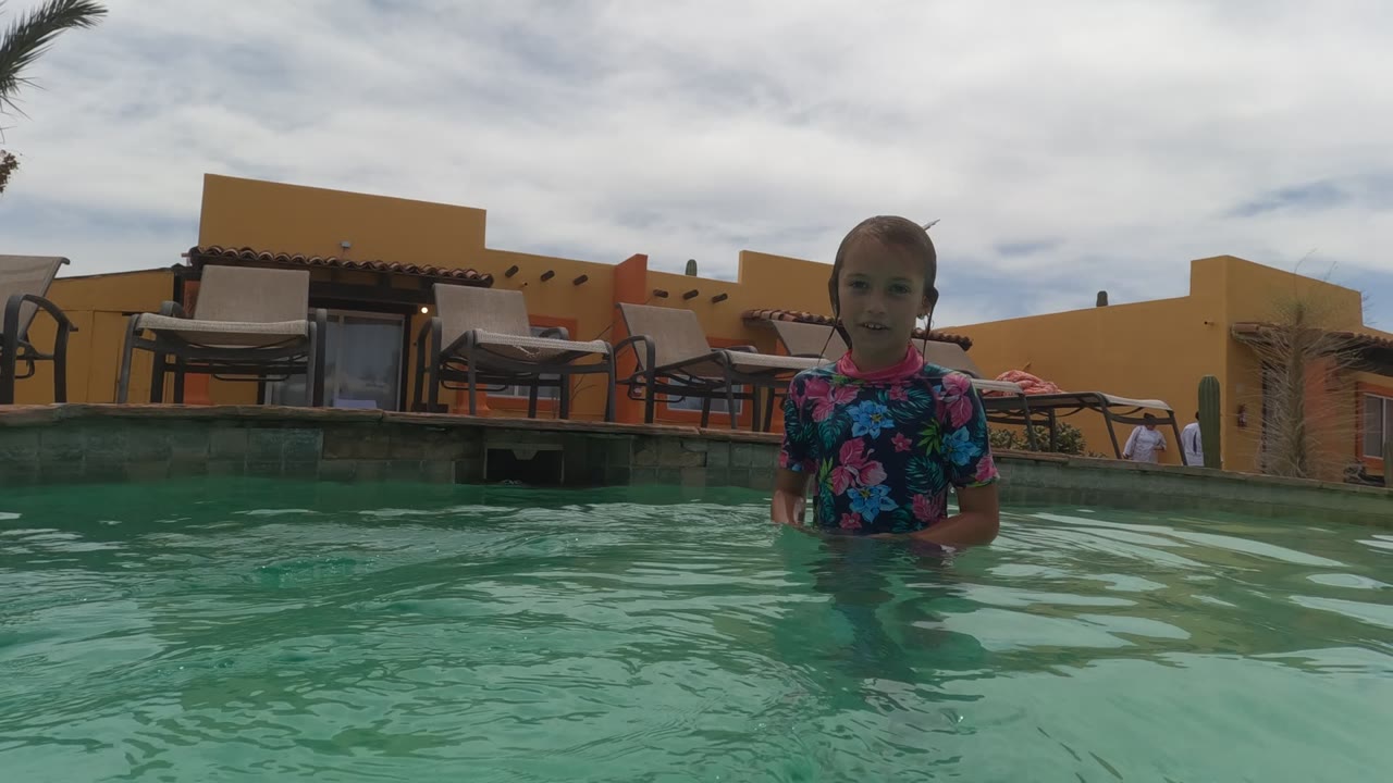 The child appears near the right side of the Baja California pool frame, partly surrounded by water, with tan lounge chairs and yellow-orange structures in the background.