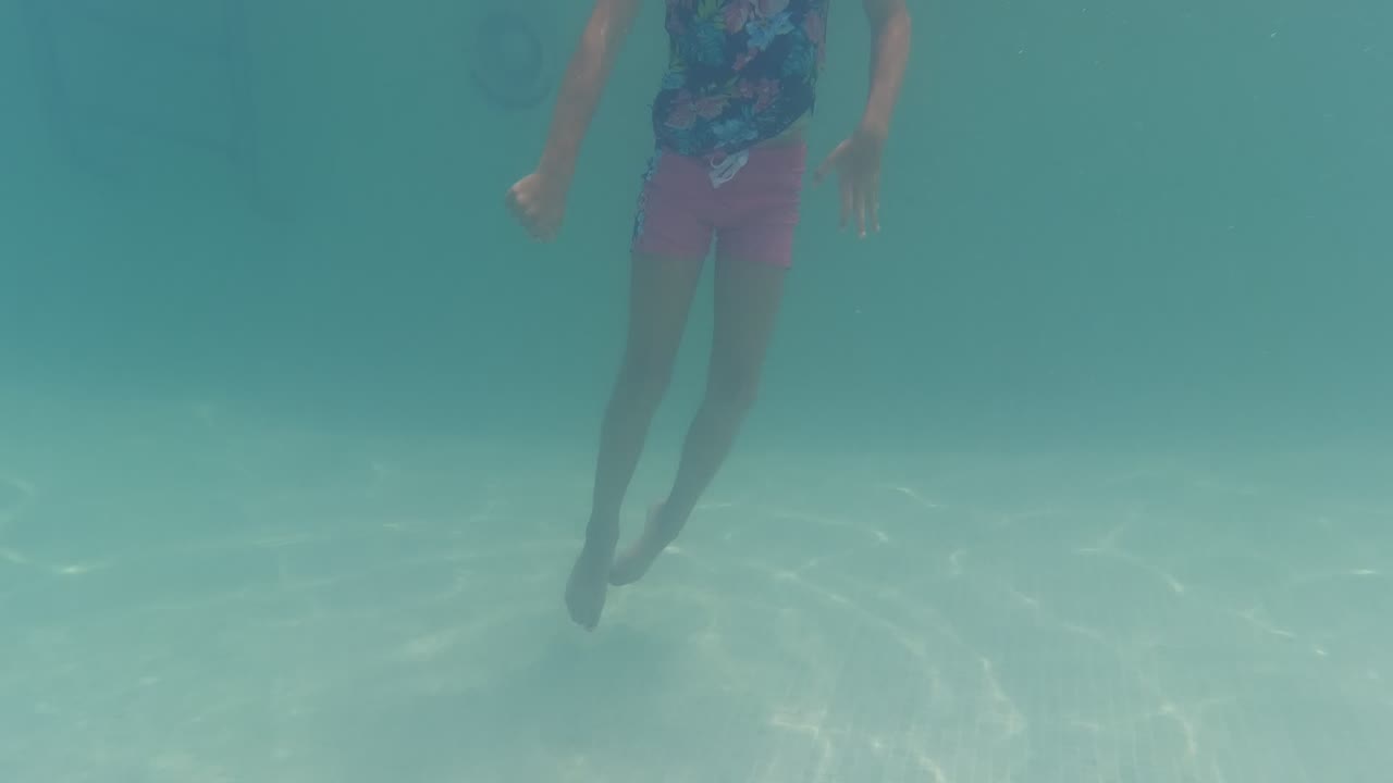 Child in a floral swim shirt and pink shorts swims underwater in a Baja California, Mexico pool with pale blue water around them.