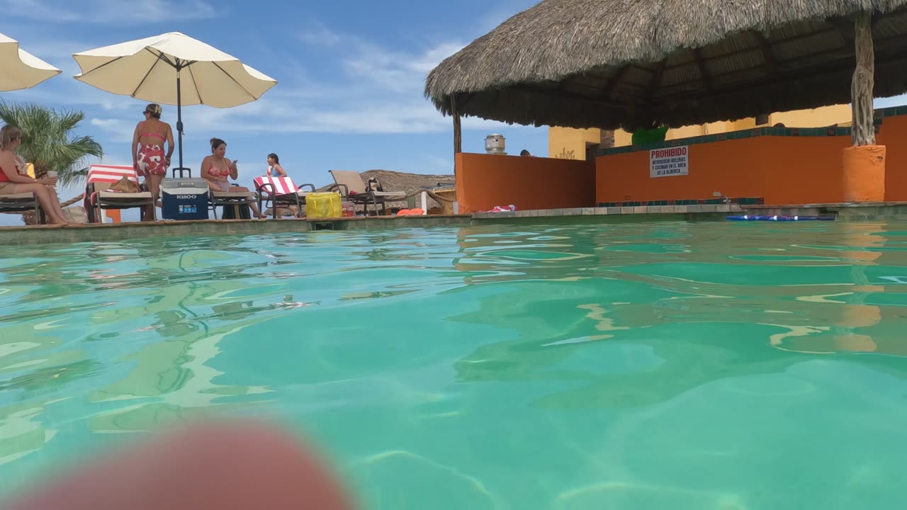 Pool-level Baja California, Mexico frame with turquoise water in front, guests near lounge chairs and umbrellas, and a thatched-roof structure behind them.