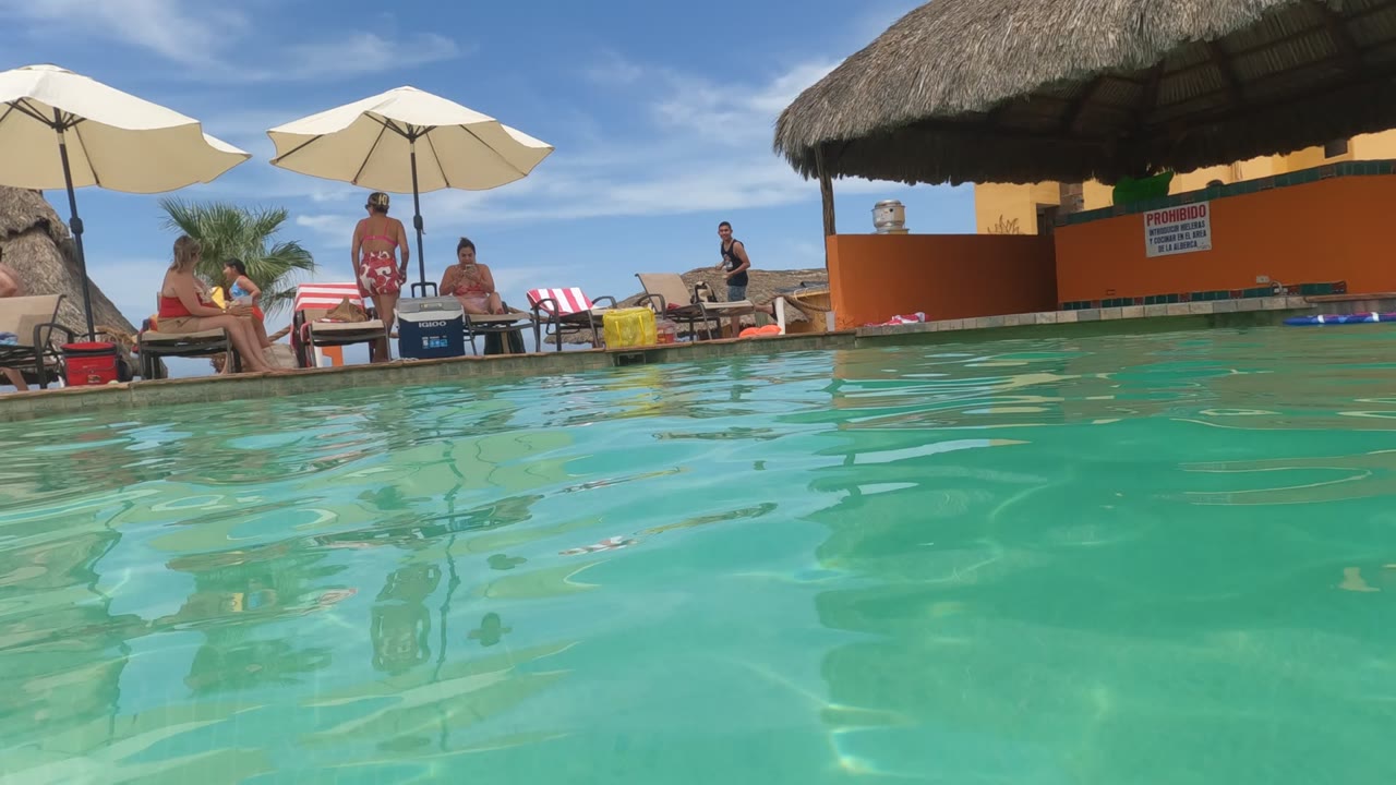 Low pool view with seated guests, umbrellas, and thatched structure.