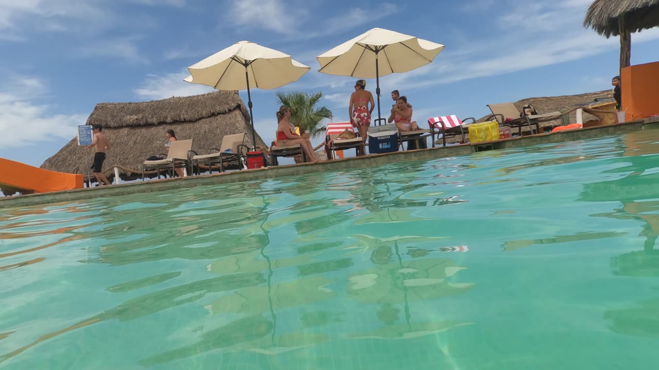 Baja California, Mexico frame with the camera tilted slightly at water level, showing clear pool water, umbrellas, lounge chairs, and people gathered near the slide area.