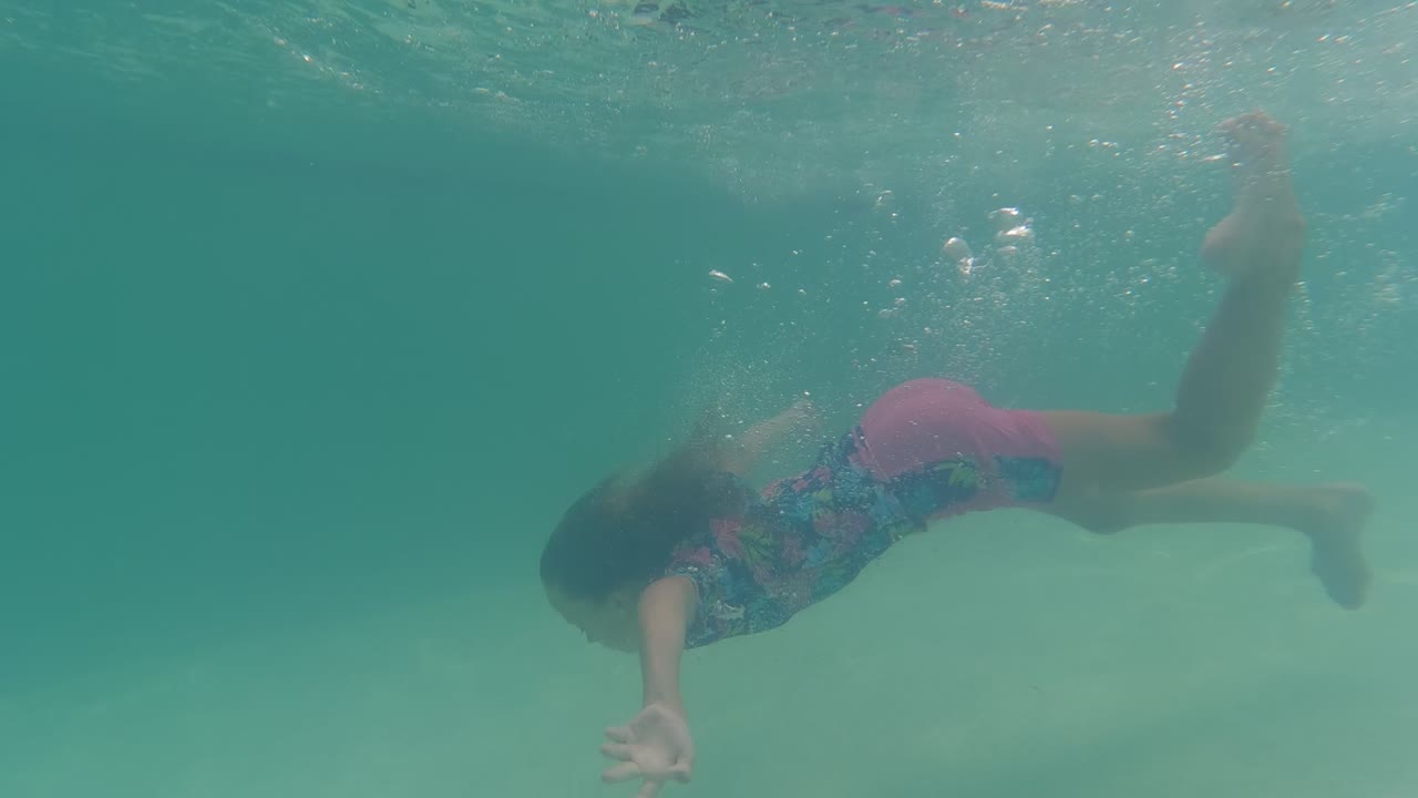 A young girl in a colorful swimsuit swims underwater in a Baja California pool, angled forward with one arm reaching down near the pale pool floor.