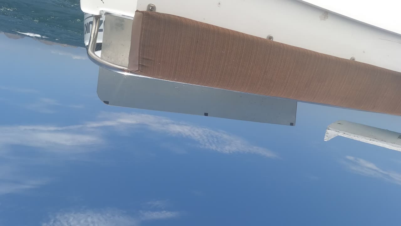 Tilted view from a yacht in Baja California, Mexico, showing a brown deck panel, white hull edge, blue ocean, scattered clouds, and no visible people.