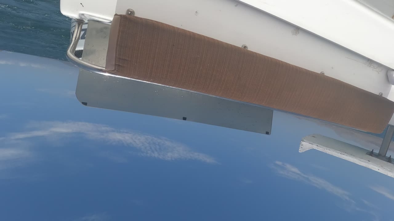 Angled Baja California yacht view with the brown deck strip crossing the foreground, white hull hardware at the edge, blue sea below, and no visible people.
