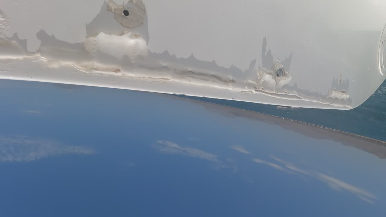 Low ship-side view in Baja California with a curved section of white hull overhead, blue sea below, scattered cloud reflections, and no visible people.