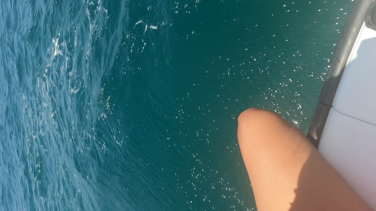 First frame of a Baja California boat ride showing blue ocean water, the white edge of the boat, and a partial leg in the foreground with no visible face.