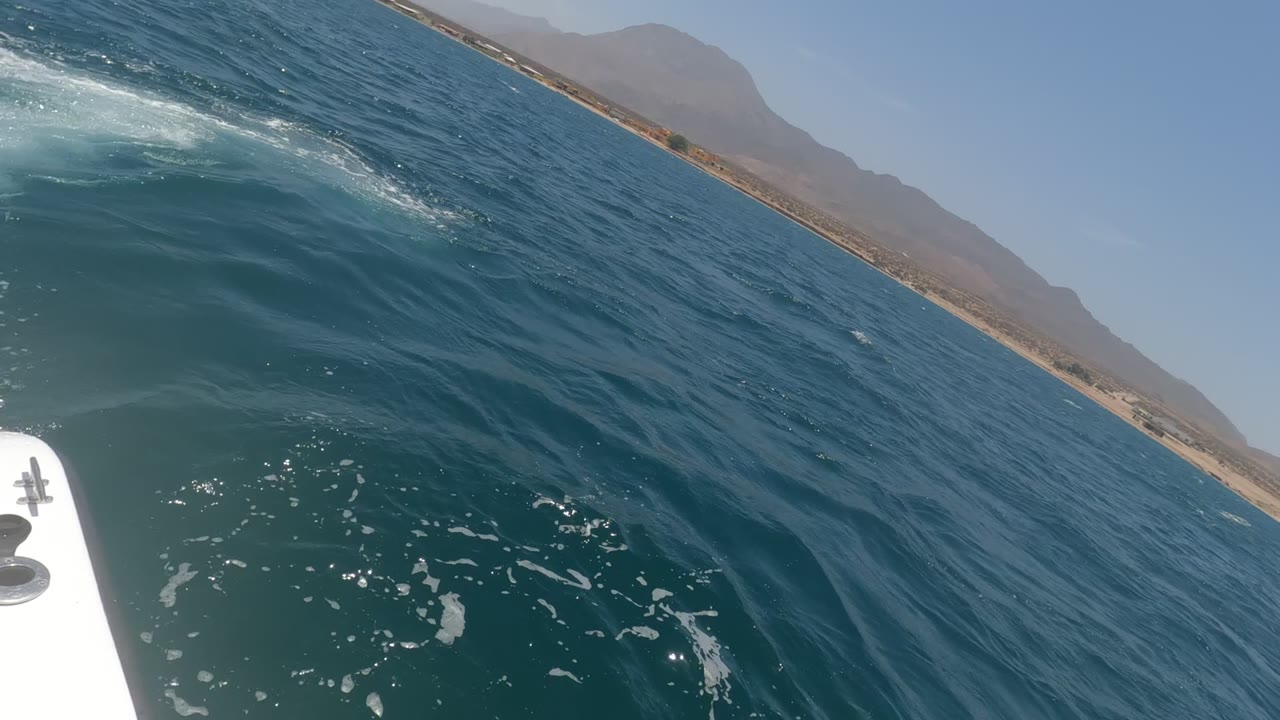 First frame of a Baja California, Mexico boat ride shows blue-green water, white wake near the left side, a white boat edge in the foreground, and distant coastline with mountains, no visible people.