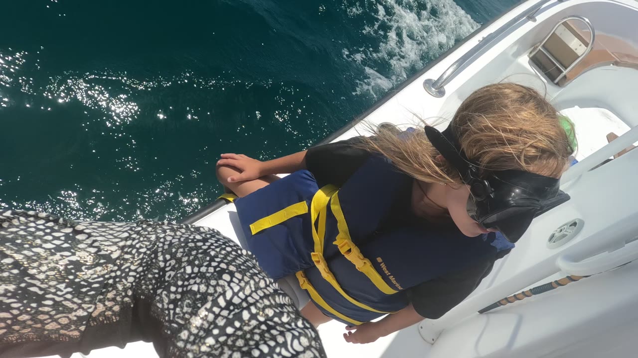 In Baja California, Mexico, a child wearing a blue and yellow life jacket and black swim mask stands on a white boat deck while teal water churns beside the boat.