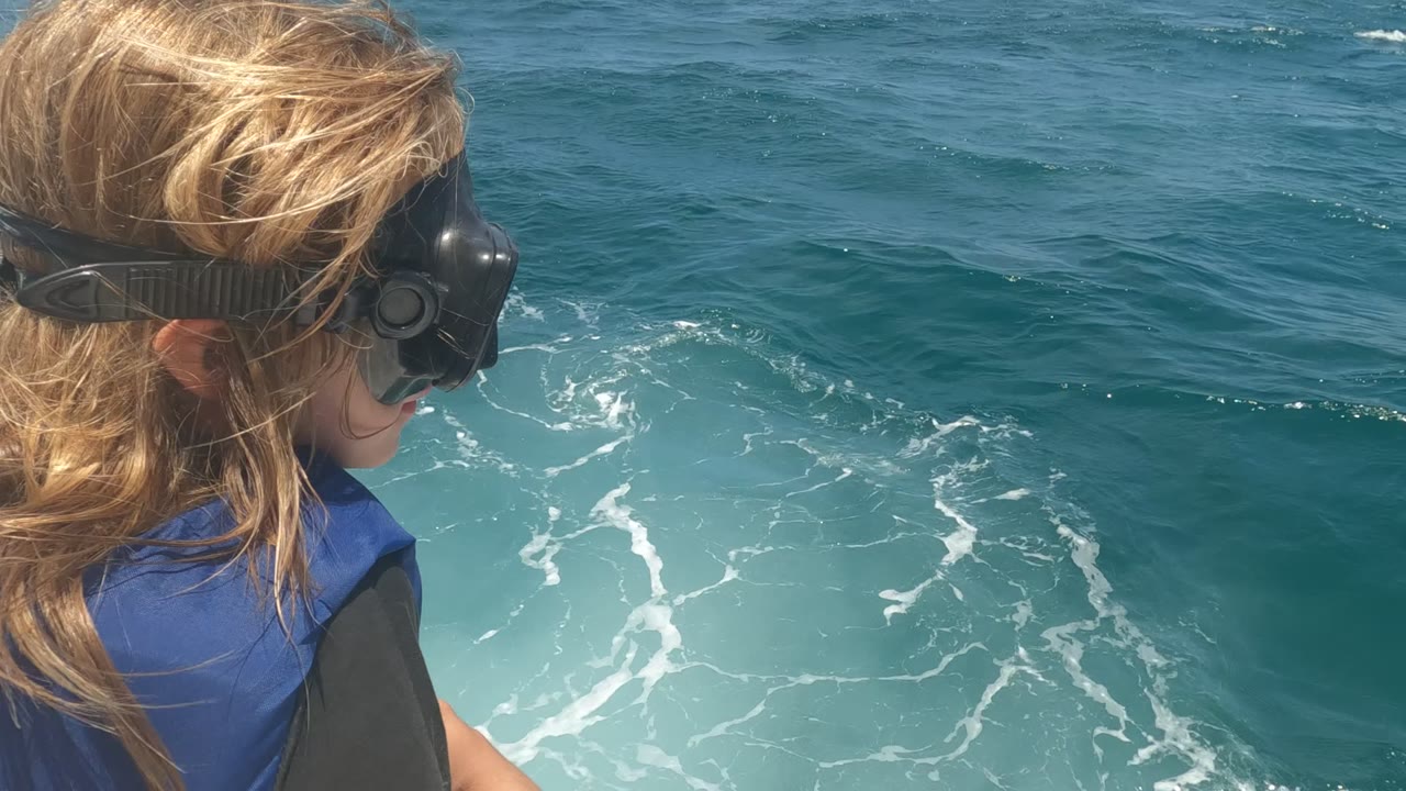 In Baja California, Mexico, the child appears closer to the right side of the frame, wearing a blue life jacket as foamy water swirls beside the boat.