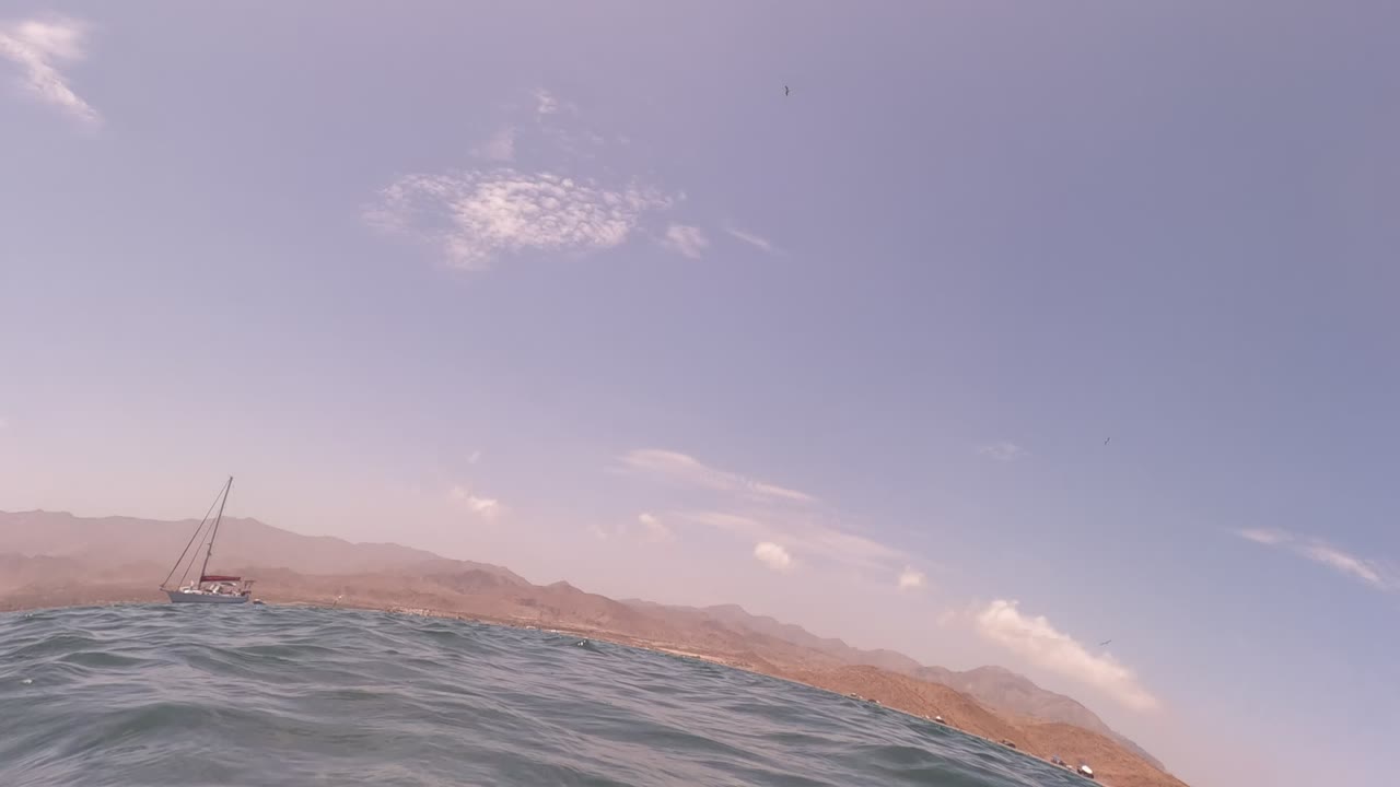 Ocean surface with a distant sailboat and brown mountains under a partly cloudy sky.
