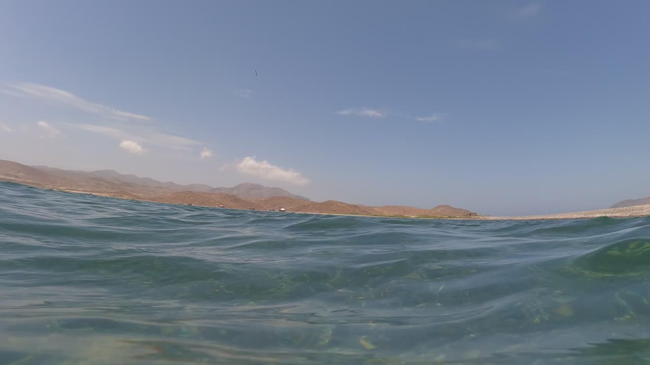 Low ocean-surface frame in Baja California, Mexico showing rounded waves, a dry mountain shoreline, blue sky, thin clouds, and no visible people.
