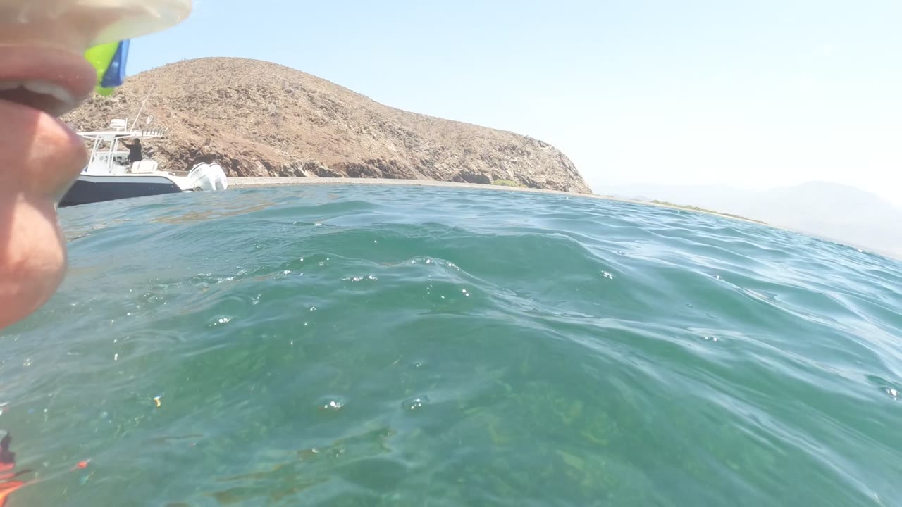 Waterline view in Baja California, Mexico with a partial snorkeler face at the left edge, green-blue ocean surface, a white boat, dry rocky coast, and distant hills.