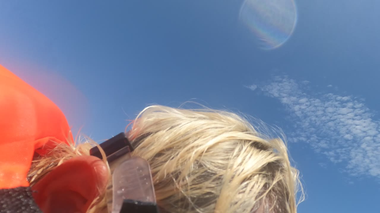 A blonde person in Baja California, Mexico adjusts snorkel gear near the edge of the frame, with orange flotation fabric and blue sky behind them.