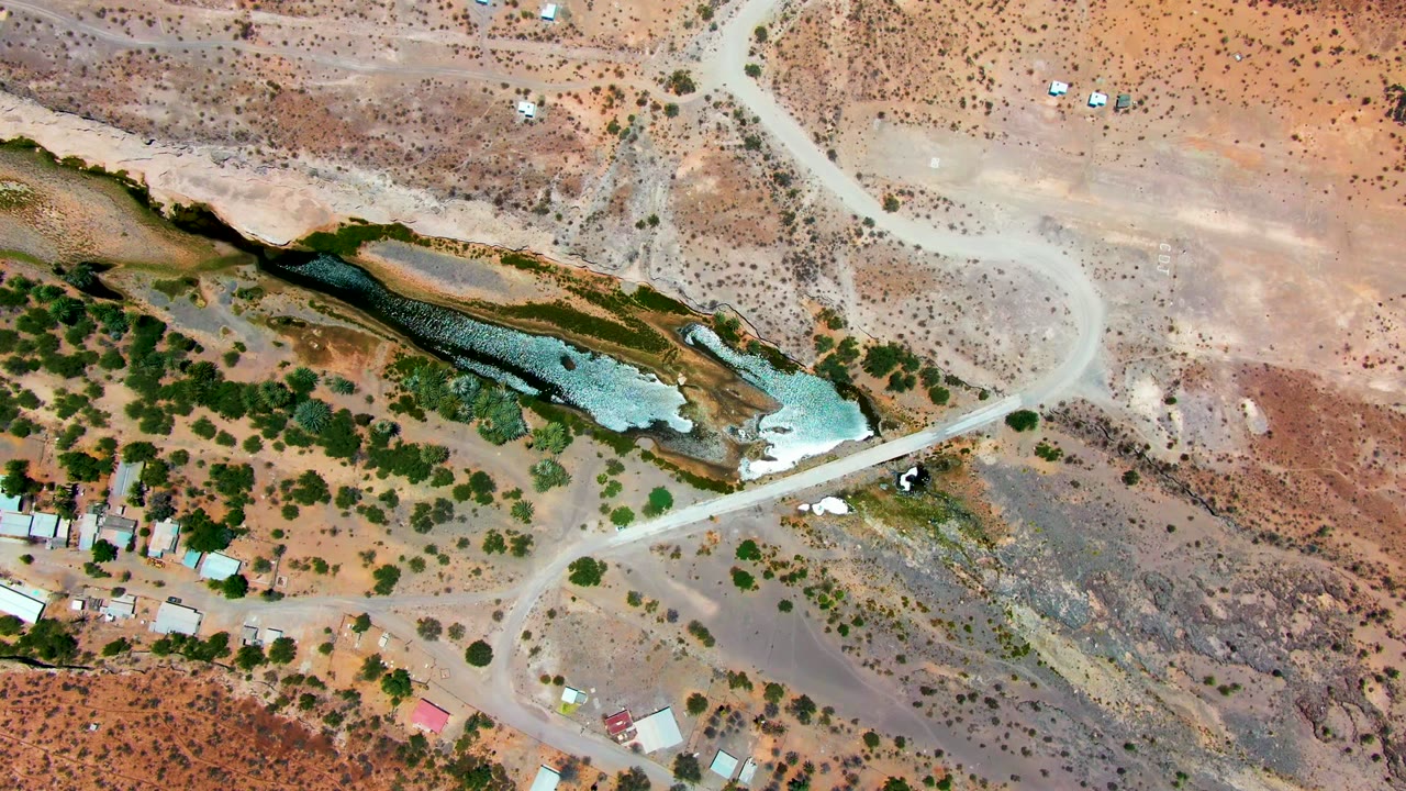 Opening overhead frame of San Juanico, Baja California Sur, with a pale dirt road crossing dry terrain beside shallow blue-green water, scattered trees, small structures, and no visible people.