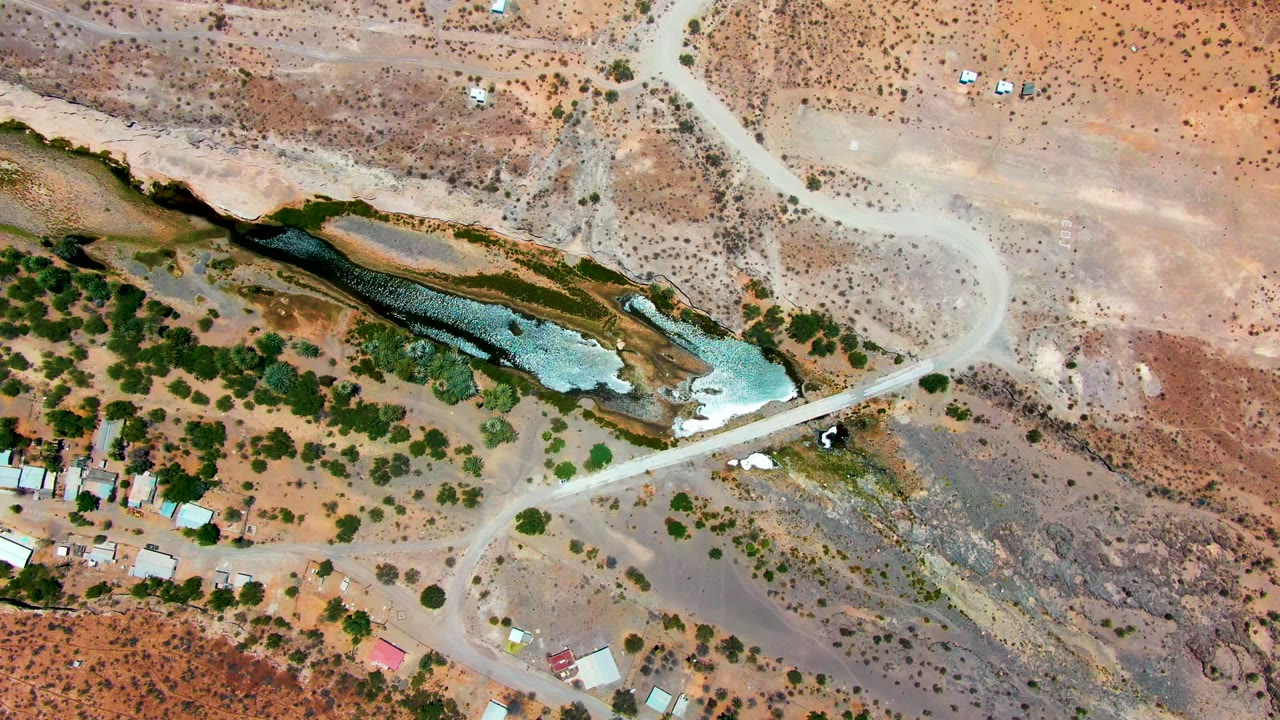 Third overhead frame of San Juanico arid terrain, where the pale road curves across the upper right and shallow water cuts through the middle beside green trees, with no visible people.