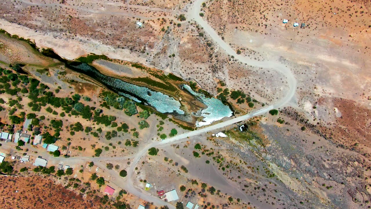 Overhead frame from San Juanico, Baja California Sur showing a winding dirt road, a green-blue water channel, scrub vegetation, scattered structures, and no visible people.