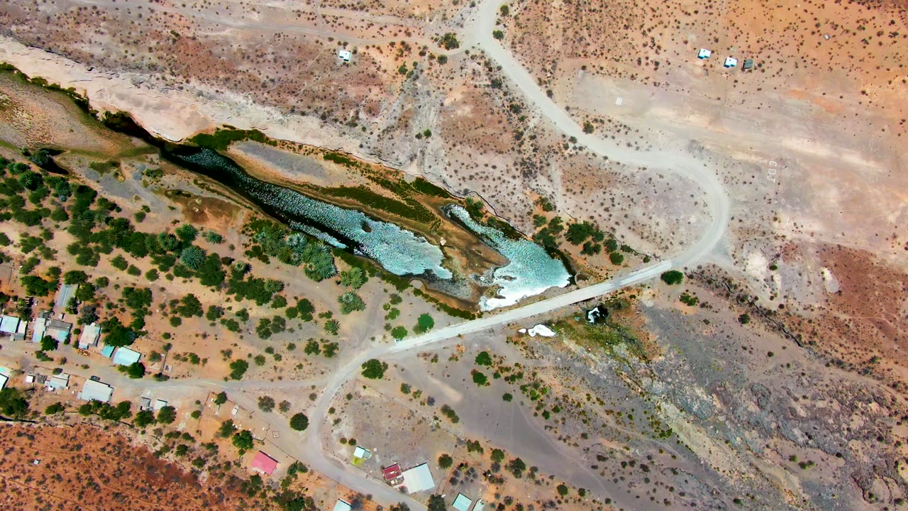 A small vehicle appears on a pale dirt road near a rocky green water body.