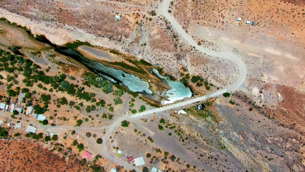 High overhead frame from San Juanico, Baja California Sur showing the road curving near the narrow green water channel, reddish scrubland, a few roofs, and no visible people.