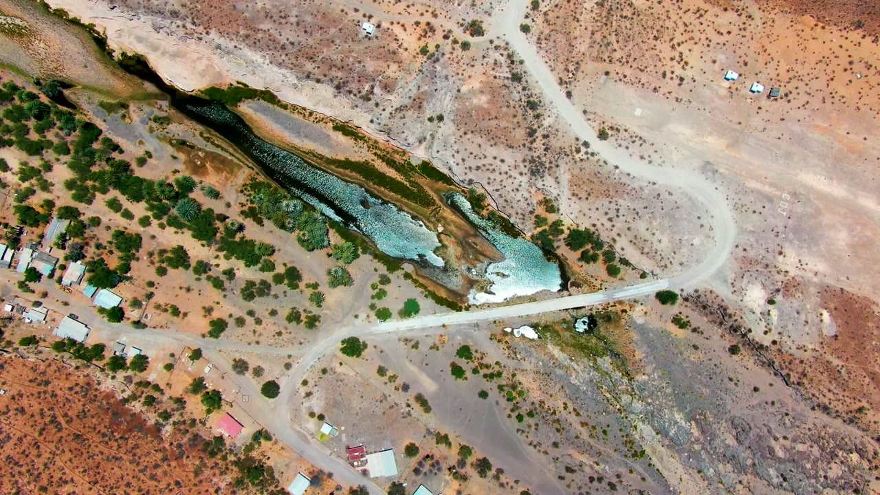 Overhead view in San Juanico, Baja California Sur showing a dirt road crossing a dry riverbed beside blue-green water, scattered trees, small buildings, and no visible people.