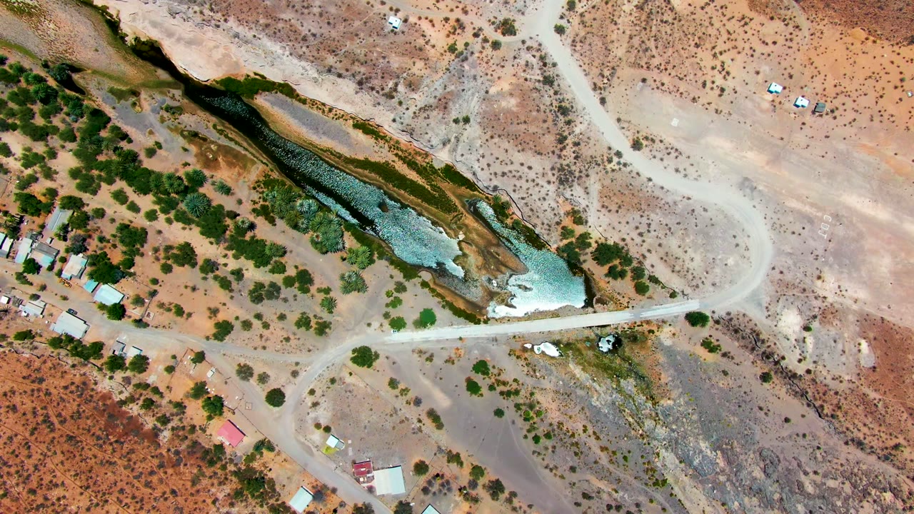 Top-down view over San Juanico, Baja California Sur where receding water cuts through the dry wash, green trees cluster nearby, dirt tracks branch across the frame, and no visible people appear.