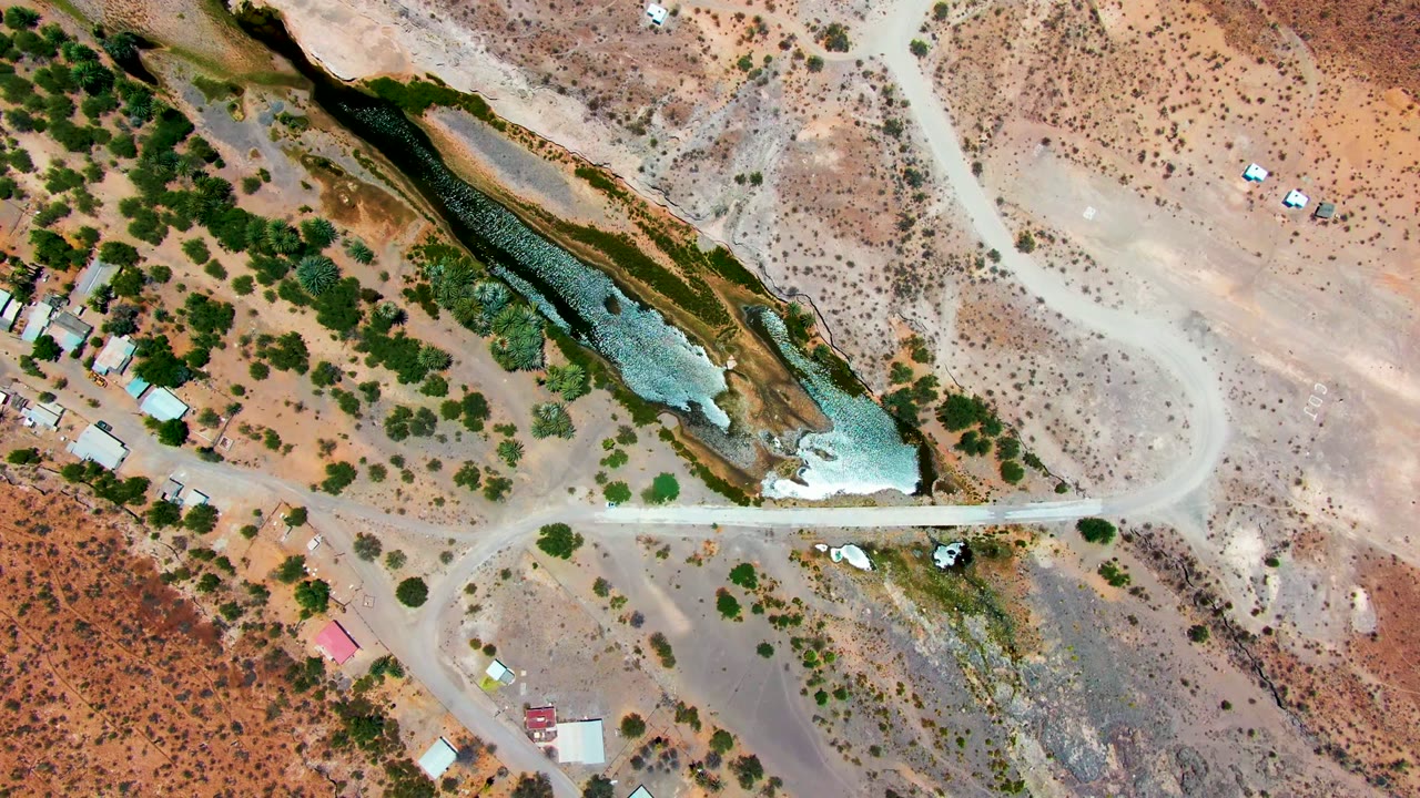 Overhead view in San Juanico, Baja California Sur showing a green-blue river channel, pale dirt road crossing, scattered shrubs, small buildings, and no visible people.