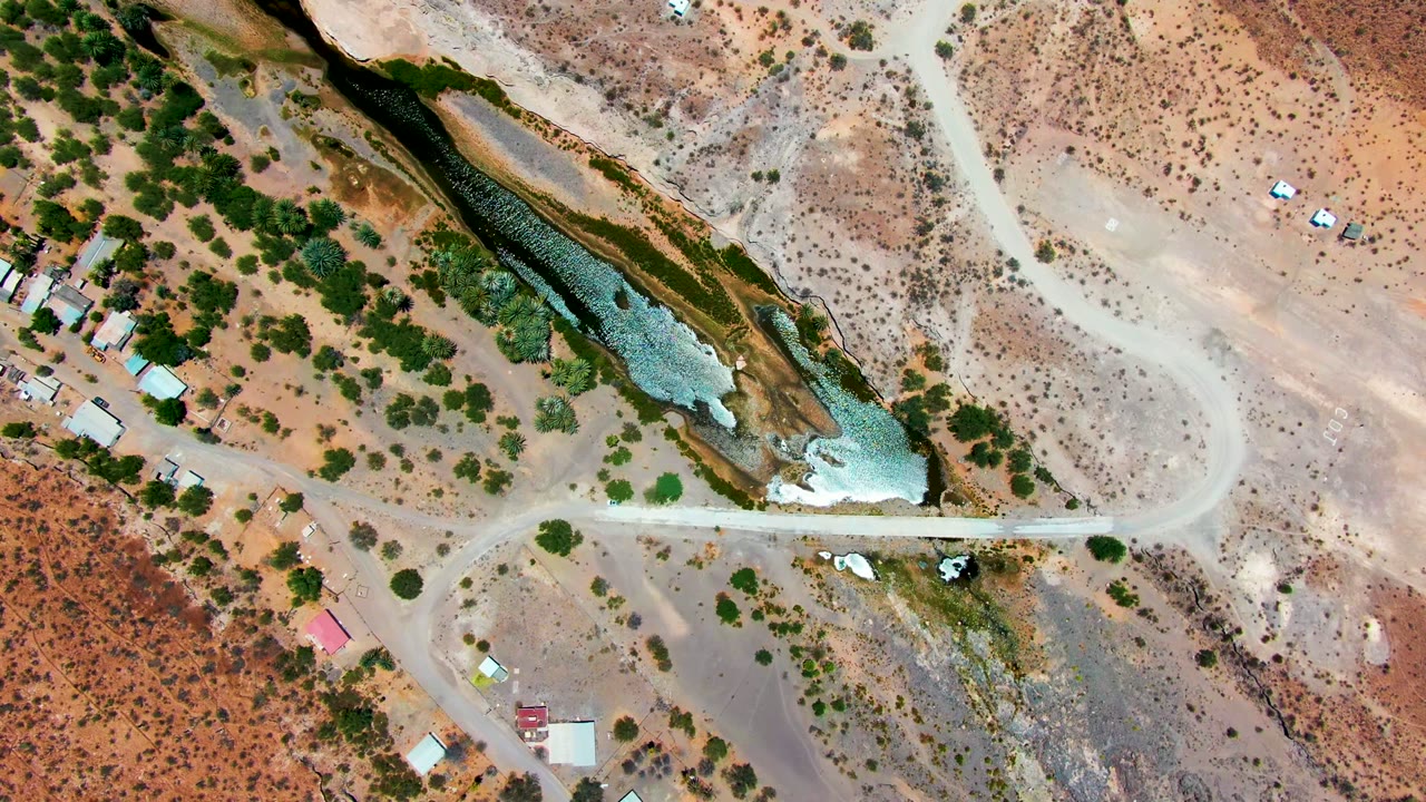 Top-down view over San Juanico, Baja California Sur showing a dark river bend, pale road tracks, rust-colored dry ground, scattered green shrubs, small buildings, and no visible people.