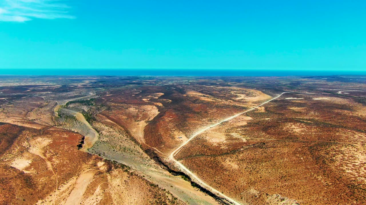 Opening aerial view over San Juanico, Baja California Sur, showing a winding riverbed beside brown rocky desert, pale dirt roads, distant horizon, and no visible people.