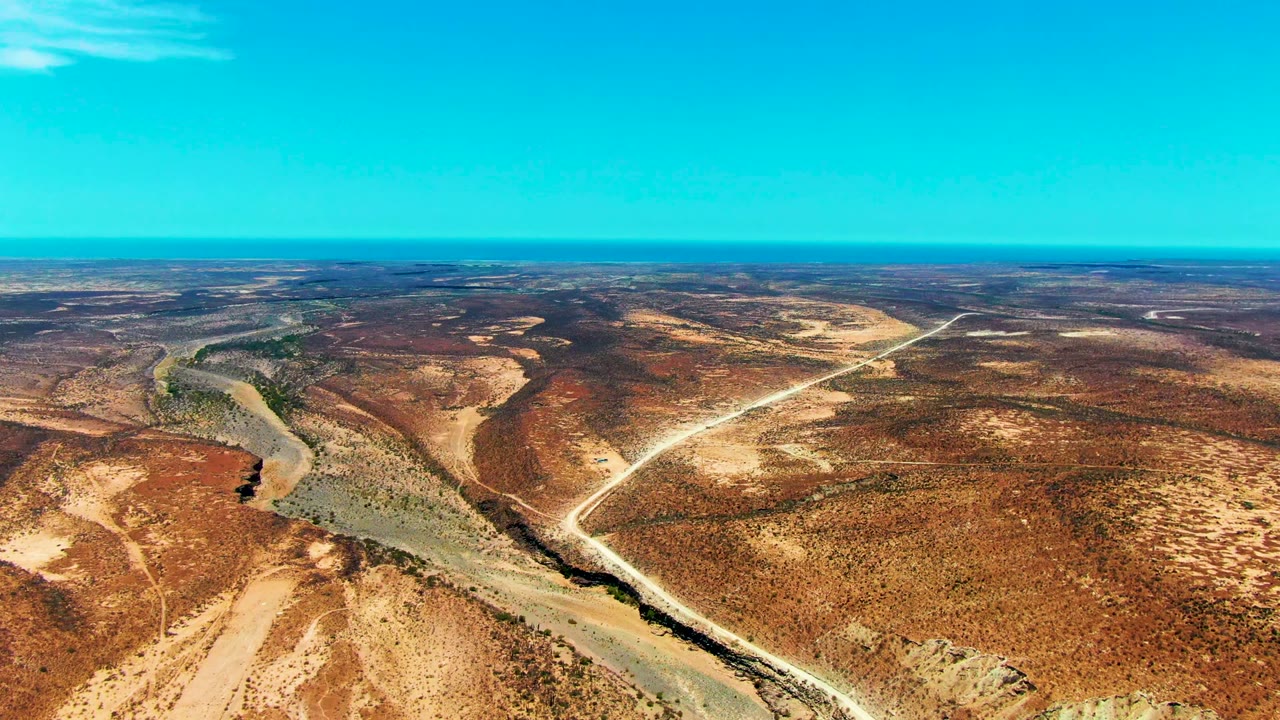 A wide aerial view over San Juanico, Baja California Sur shows a winding river at left, pale dirt road cuts, brown arid terrain, and no visible people.