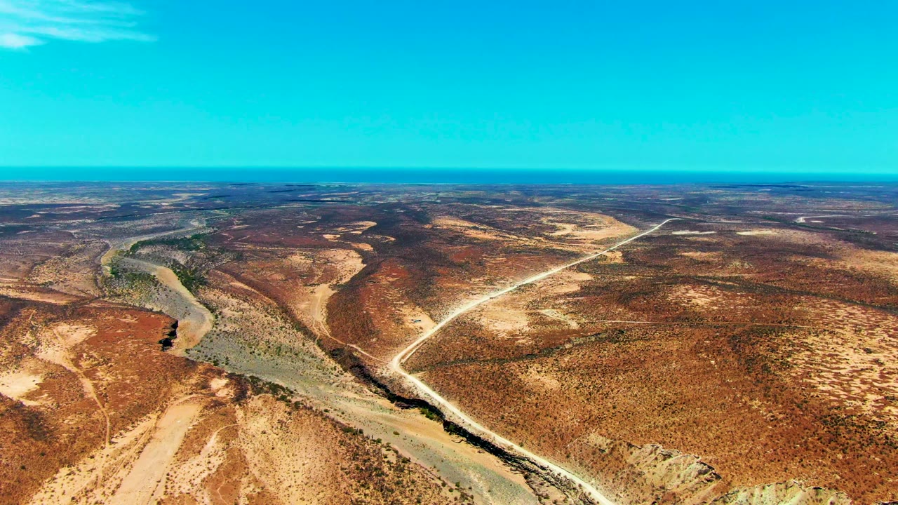 Wide aerial desert plain with river bend, road tracks, sparse vegetation, and bright sky.