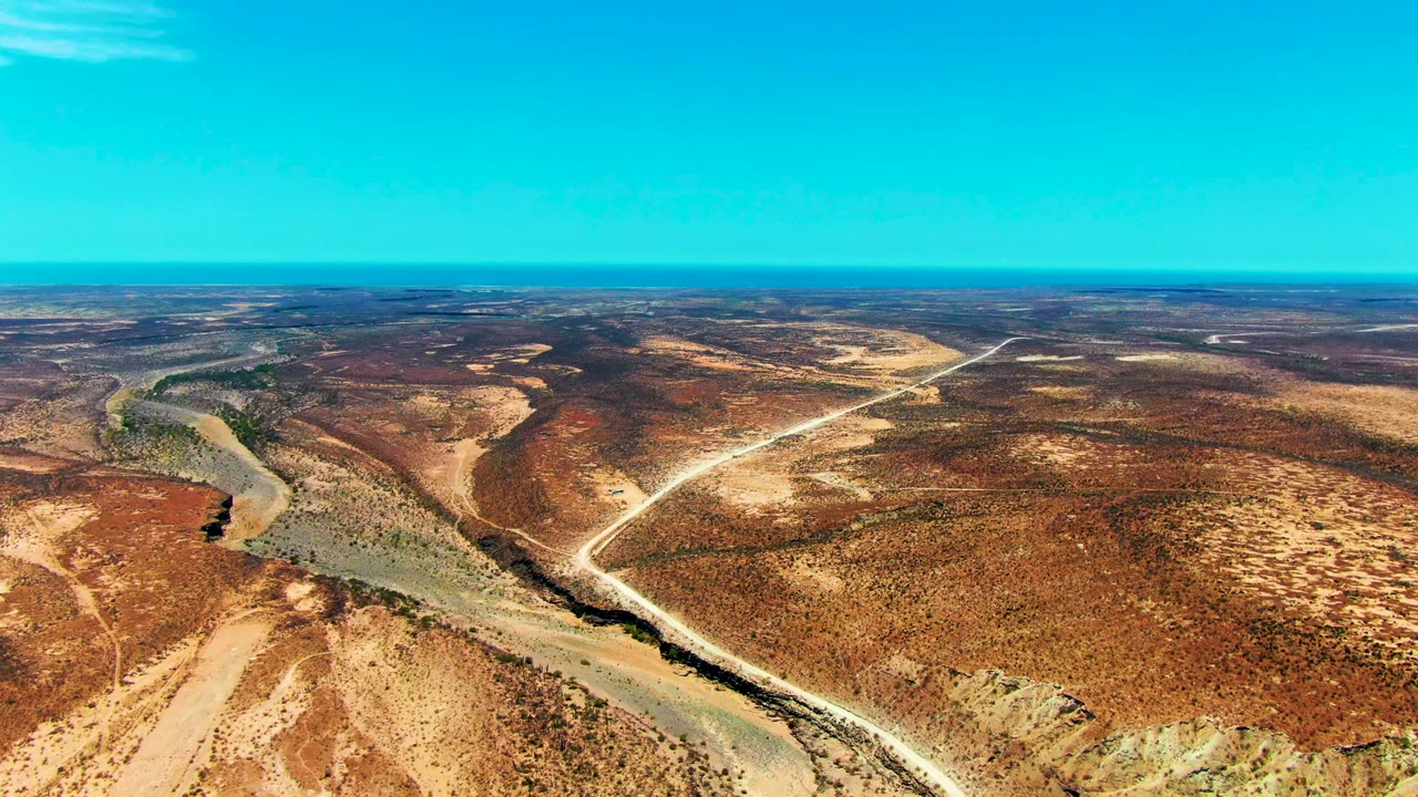 Opening aerial frame over San Juanico, Baja California Sur, with a green-gray river bending through brown desert scrub, pale dirt roads crossing the terrain, a flat horizon, and no visible people.